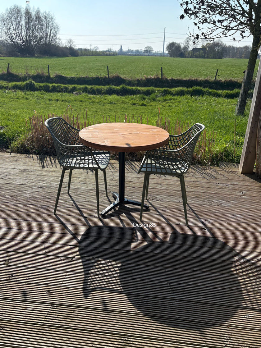 Kleine ronde terrastafel met zwart metalen poot en houten blad, geplaatst tussen twee groene kunststof stoelen in een zonnige tuin.