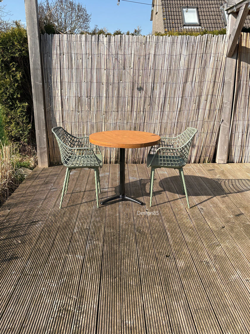Buitenopstelling met ronde houten horecatafel, twee groene stoelen en een sfeervolle tuinsetting met schutting en zonlicht.