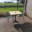 Industriële bartafel met houten blad op een terras in de tuin, omringd door planten en moderne stoelen.