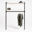 Industriële kledingkast van zwart staal met twee legplanken van onbehandeld vurenhout, op een witte achtergrond met hangende T-shirts, boots en pet.