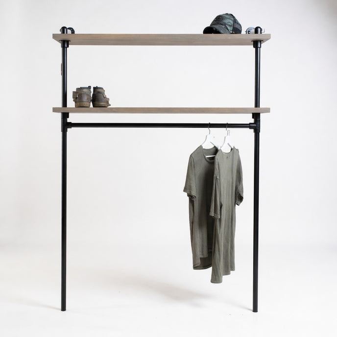 Industriële kledingkast van zwart staal met twee legplanken van vurenhout in taupe wax, op een witte achtergrond met hangende T-shirts, boots en pet.